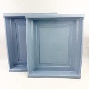 May include: Two rectangular, light blue plastic trays with a smooth, glossy finish. Each tray features a raised border and a recessed central panel, creating a clean, structured design. The trays are stacked slightly offset from one another.