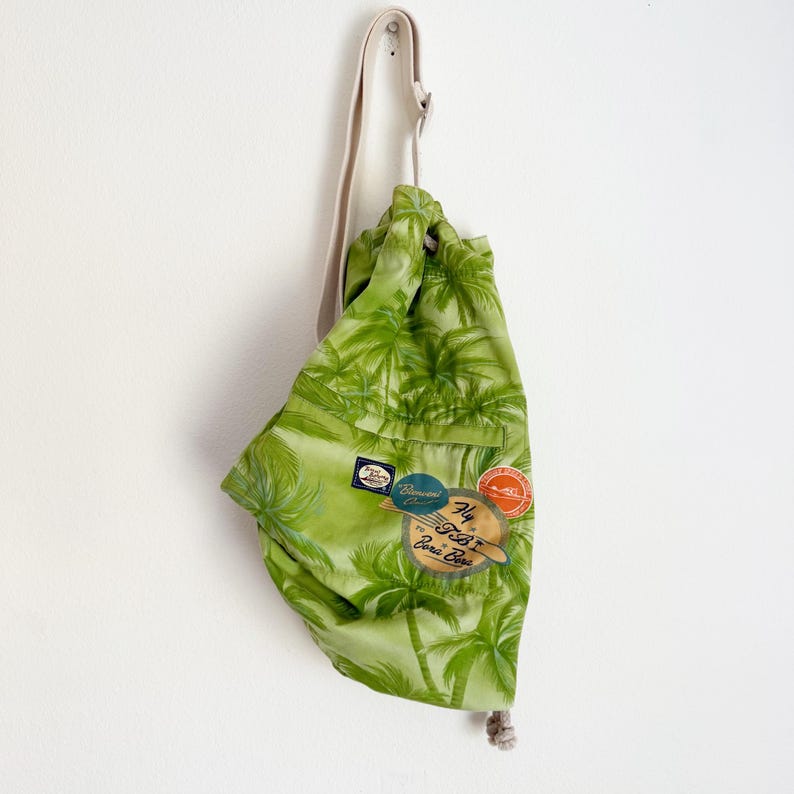 May include: A green drawstring backpack with a tropical palm tree pattern. The bag has a beige shoulder strap and several colorful patches. The text "Fly Bora Bora" is visible on one of the patches.