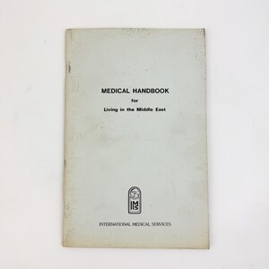 May include: A white softcover book titled Medical Handbook for Living in the Middle East. The cover features black text, a small circular logo with a globe icon above the letters IMS, and the text International Medical Services at the bottom.