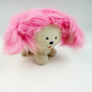 May include: A plush toy dog with a cream-colored body and a large, fluffy pink mane. The dog has black eyes, a blue nose, and a red tongue sticking out. The toy is sitting on a white surface.