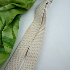 May include: A green and white patterned fabric with palm trees is next to a cream-colored canvas strap. The strap is attached to a metal buckle. The fabric is a bright green color.