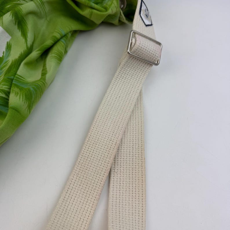 May include: Close-up of a green and white patterned fabric, possibly a bag or accessory, with a cream-colored canvas strap. The strap features a silver metal buckle and a small tag.