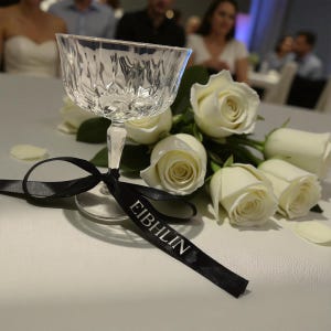 May include: A crystal champagne coupe with a black ribbon, the word "EIBHLIN" printed on it, rests on a white surface. White roses are arranged beside the glass. The coupe has a detailed, textured design.