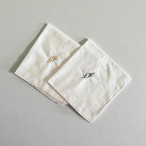 May include: Two white handkerchiefs with embroidered initials. One handkerchief has gold-colored initials, and the other has black initials. Both handkerchiefs have a striped pattern.