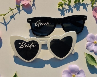 May include: Two pairs of sunglasses, one black and one white. The black sunglasses have the word "Groom" in white script. The white sunglasses are heart-shaped and have the word "Bride" in white script. The sunglasses are surrounded by flowers and greenery.