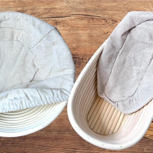 Peut inclure: Deux paniers à lever blancs avec des doublures en lin. Un panier est rond et l'autre est ovale. Le panier ovale est à côté d'un pain et d'épis de blé.