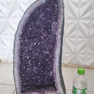 May include: A large amethyst geode with a dark grey base and a deep purple crystal interior. The geode is next to a clear plastic water bottle with a green cap. The background is a white geometric wall.