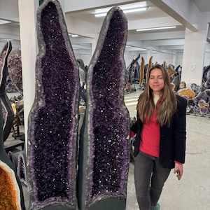 May include: Two large, tall amethyst geodes standing upright, featuring deep purple, crystalline interiors with jagged, sparkling surfaces. The outer edges of the geodes are dark, rough, and rock-like, contrasting with the vibrant, faceted purple crystals inside.