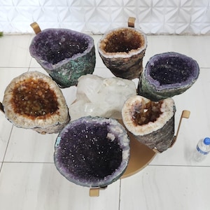 May include: A collection of amethyst and quartz geodes arranged on a gold-colored table. The geodes display shades of purple, brown, and white, with rough, textured exteriors. The table has curved metal supports.
