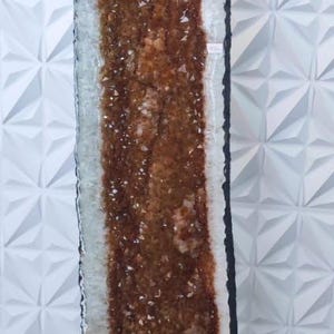 Op de afbeelding: Een grote, rechthoekige geode met een donkerbruin kristallijn centrum en een witte en heldere buitenlaag. De geode staat tegen een witte, getextureerde muur. De randen zijn zwart.