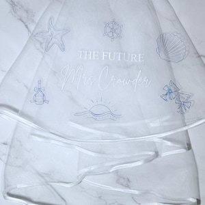 May include: White bridal veil with blue embroidered nautical themed designs, including a starfish, a ship's wheel, a seashell, a champagne bottle with a bow, a sunrise over the ocean, and two champagne flutes with bows. The text "THE FUTURE Mrs. Crowder" is embroidered on the veil.