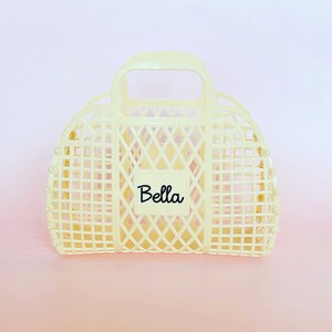 May include: A cream-colored plastic tote bag with a handle and a diamond-shaped grid pattern. The bag has a label that says "Bella" on it.