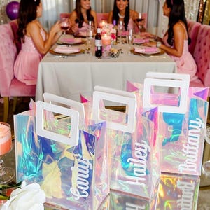 May include: Three rectangular iridescent gift bags with white plastic handles. Each bag features a custom white script name decal on the front, reading Carly, Hailey, and Brittany, reflecting light with a holographic, multi-colored sheen.