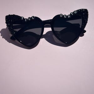 May include: Black heart-shaped sunglasses featuring dark lenses and black glitter embellishments on the outer frames. The top of the frames displays white letter beads spelling out the phrase TILL DEATH across the two heart-shaped rims.