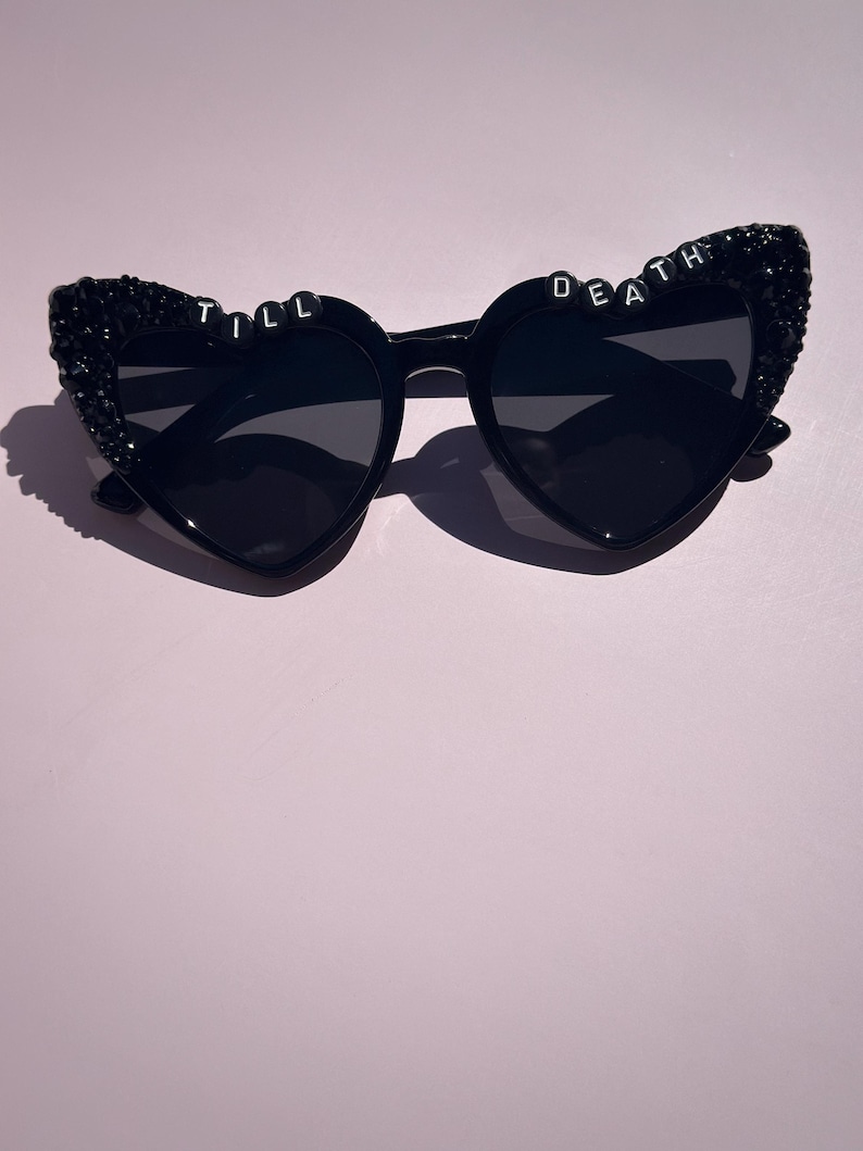 May include: Black heart-shaped sunglasses featuring dark lenses and black glitter embellishments on the outer frames. The top of the frames displays white letter beads spelling out the phrase TILL DEATH across the two heart-shaped rims.