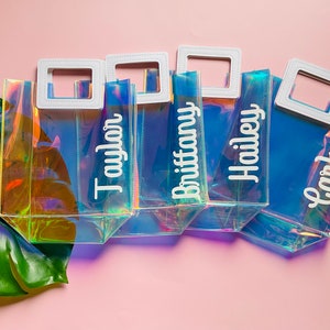K&ouml;nnte beinhalten: Vier durchsichtige, regenbogenfarbene Plastikt&uuml;ten mit wei&szlig;en Griffen und wei&szlig;en Beschriftungen. Die Taschen sind mit "Taylor", "Brittany", "Hailey" und "Carly" beschriftet.