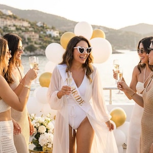 May include: A white sash with the word BRIDE printed in gold lettering, worn over a white swimsuit and a sheer white cover-up. The person is wearing white heart-shaped sunglasses and holding a glass of champagne on a boat.