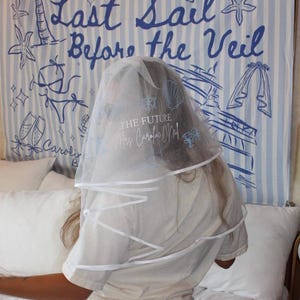 May include: A white sheer bridal veil featuring a white ribbon trim and printed blue graphics including a seashell, a ship wheel, and two champagne glasses. The text on the veil reads THE FUTURE Mrs Caroline O'Neil in a mix of fonts.