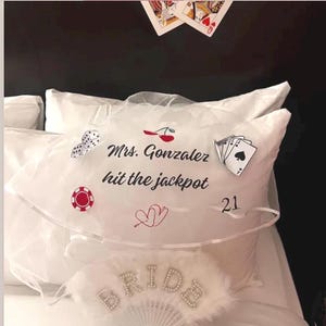 May include: A white bridal veil featuring the black text Mrs. Gonzalez hit the jackpot, decorated with red cherries, white dice, a red poker chip, playing cards, the number 21, and two red hearts. Below, a white feathered fan displays the word BRIDE.