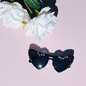 May include: Black heart-shaped sunglasses featuring a glittery finish on the frames and the text TILL DEATH spelled out in white letter beads across the top edges of the lenses, accompanied by two white artificial roses with green stems.