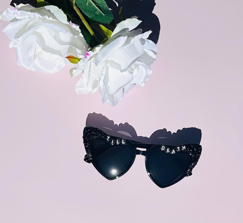 May include: Black heart-shaped sunglasses featuring a glittery finish on the frames and the text TILL DEATH spelled out in white letter beads across the top edges of the lenses, accompanied by two white artificial roses with green stems.