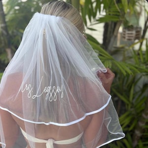 May include: A white, two-tier tulle bridal veil featuring a satin ribbon trim along the bottom edge. The veil is embroidered with the white cursive text "Mrs. Leggsey" on the lower section of the fabric, creating a personalized decorative accent.