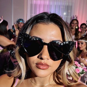 May include: Black heart-shaped sunglasses featuring decorative black embellishments on the outer corners and the text TILL DEATH spelled out in white letters across the top of the frames. The lenses are dark and opaque, providing a bold look.