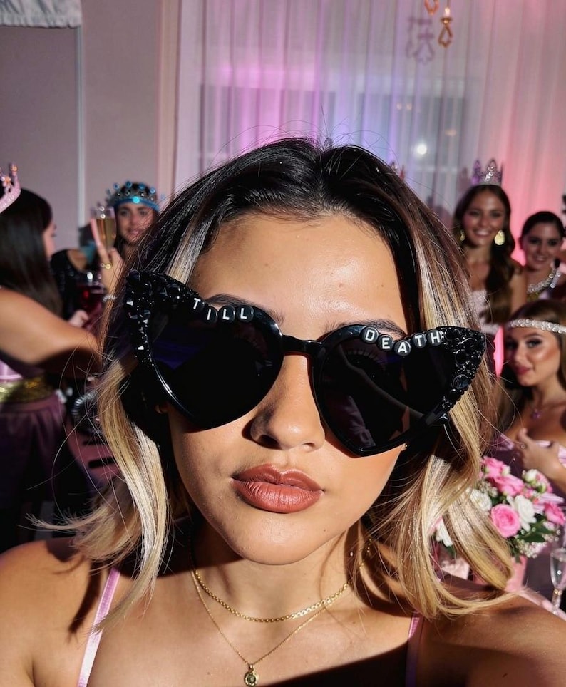 May include: Black heart-shaped sunglasses featuring decorative black embellishments on the outer corners and the text TILL DEATH spelled out in white letters across the top of the frames. The lenses are dark and opaque, providing a bold look.