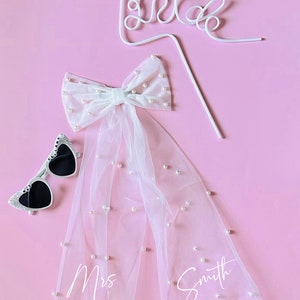 May include: A white tulle veil with pearl embellishments and the text "Mrs. Smith" written on it. A white bow with pearls is attached to the top of the veil. A white wire "Bride" sign and a pair of heart-shaped sunglasses are also included.