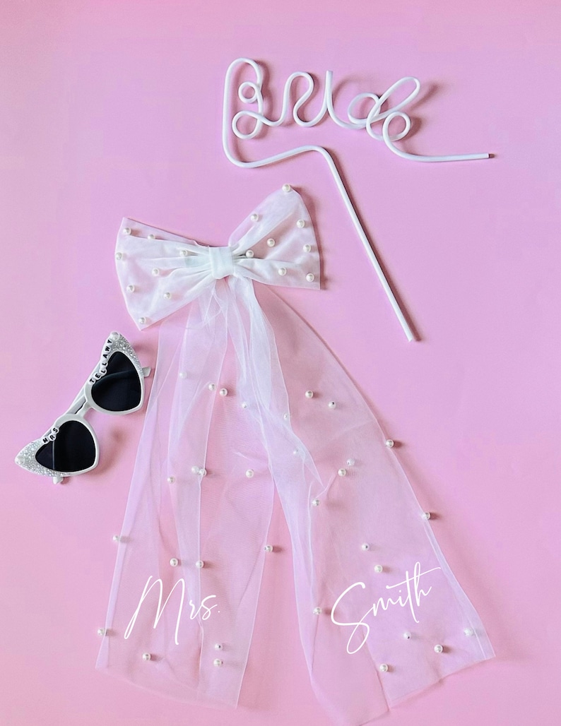 May include: A white tulle veil with pearl embellishments and the text "Mrs. Smith" written on it. A white bow with pearls is attached to the top of the veil. A white wire "Bride" sign and a pair of heart-shaped sunglasses are also included.