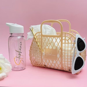 May include: A pink plastic tote bag with the name "Stephanie" written on it. The bag is filled with a white cloth and a pair of white sunglasses. A clear water bottle with a white lid and the name "Stephanie" written on it is next to the bag.