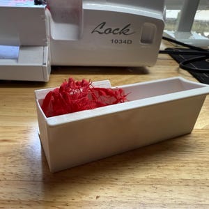 May include: A white plastic sewing machine accessory with a red lace fabric inside. The accessory is labeled "Lock 1034D".