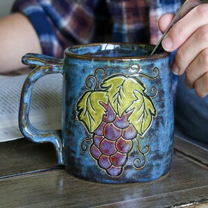 May include: A blue ceramic mug with a grape design. The mug has a textured surface and a curved handle. The grape design features green leaves and purple grapes. The mug is sitting on a wooden surface.