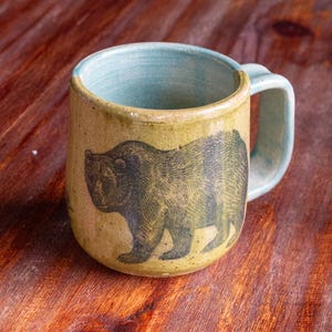 May include: A handmade ceramic mug with a bear illustration. The mug has a light green exterior and a blue interior and handle. The bear is depicted in a dark green color. The mug is sitting on a wooden surface.