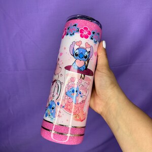 May include: A pink glitter tumbler featuring a cartoon character design. The tumbler has a white background with pink hearts and floral accents. The design includes a blue cartoon character with heart-shaped details. The tumbler is held by a hand.