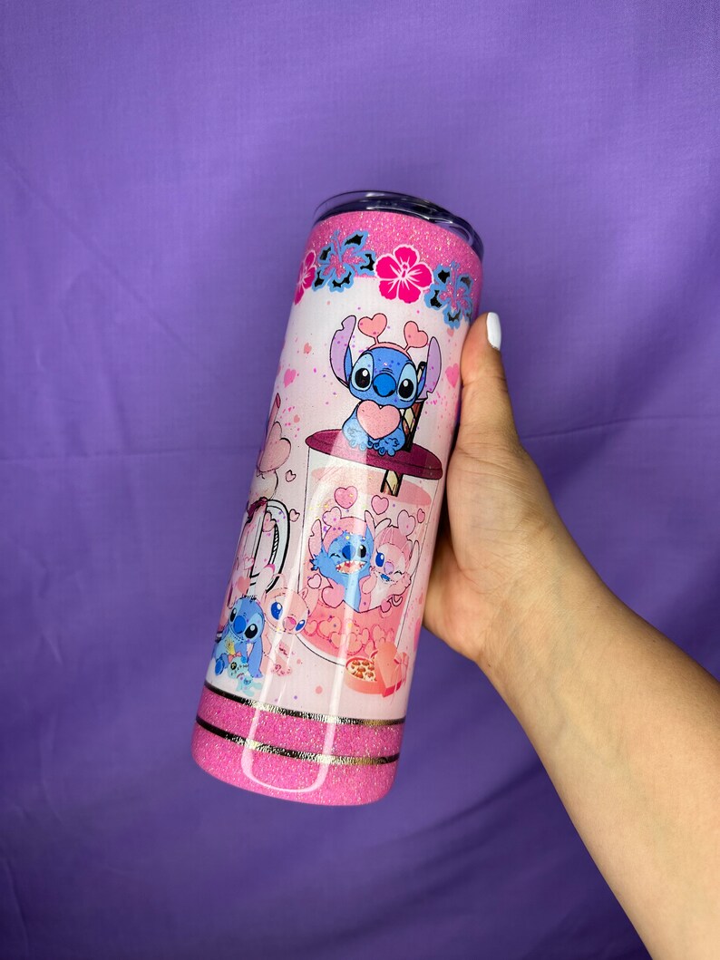 May include: A pink glitter tumbler featuring a cartoon character design. The tumbler has a white background with pink hearts and floral accents. The design includes a blue cartoon character with heart-shaped details. The tumbler is held by a hand.