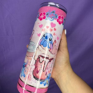 May include: A pink glitter tumbler featuring a cartoon character design. The tumbler has a pink glitter top and bottom, with a gold band separating the two. The design includes hearts, flowers, and the character in a milkshake.