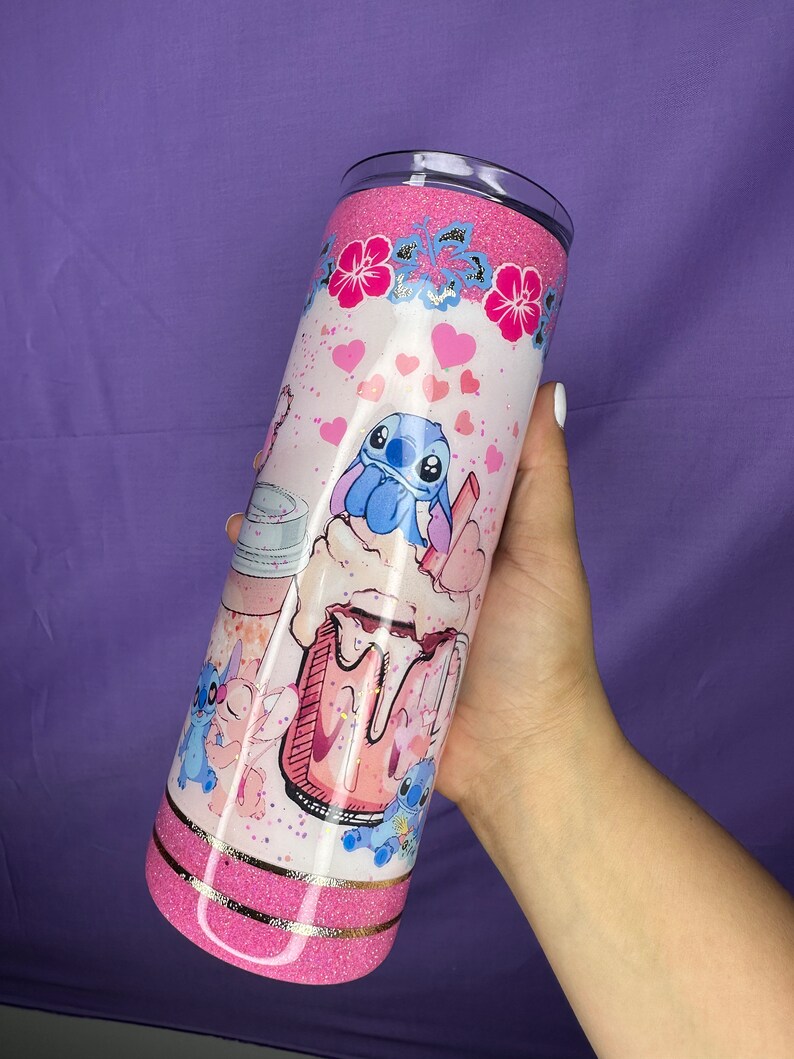 May include: A pink glitter tumbler featuring a cartoon character design. The tumbler has a pink glitter top and bottom, with a gold band separating the two. The design includes hearts, flowers, and the character in a milkshake.