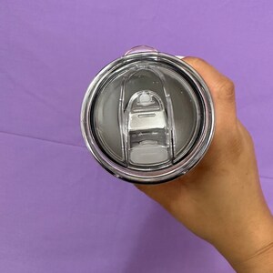 May include: A stainless steel tumbler lid with a clear plastic sliding closure. The lid is black and silver, with a clear plastic slider to open and close the drinking hole. The tumbler is held against a purple background.