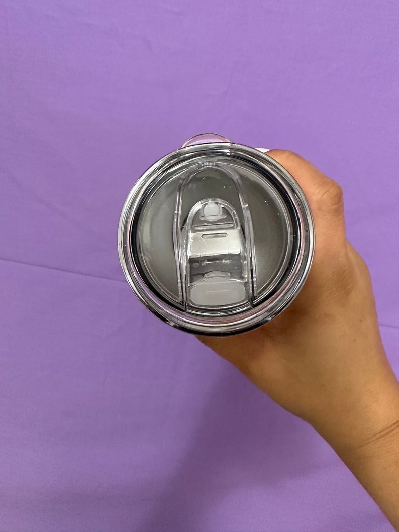 May include: A stainless steel tumbler lid with a clear plastic sliding closure. The lid is black and silver, with a clear plastic slider to open and close the drinking hole. The tumbler is held against a purple background.