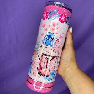 May include: A glittery pink tumbler featuring a cartoon Stitch design. The tumbler has a pink glitter top and bottom, with a gold band separating the two. The design includes hearts, flowers, and a cartoon image of Stitch enjoying a drink.