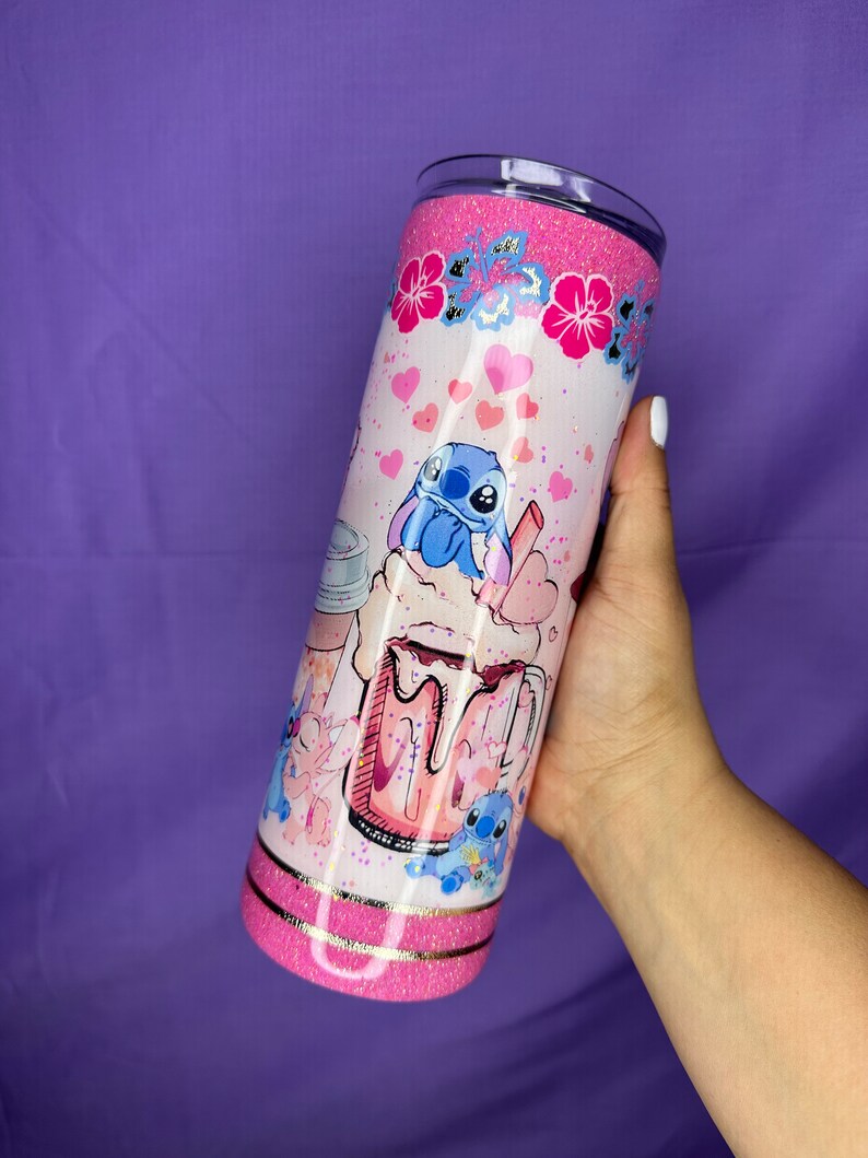 May include: A glittery pink tumbler featuring a cartoon Stitch design. The tumbler has a pink glitter top and bottom, with a gold band separating the two. The design includes hearts, flowers, and a cartoon image of Stitch enjoying a drink.