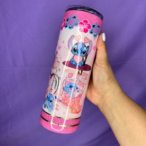 May include: A pink and white tumbler featuring a cartoon character design. The tumbler has a glittery pink top and bottom, with a design of the character holding a heart. The design includes pink hearts and flowers.