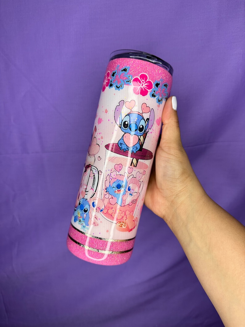 May include: A pink and white tumbler featuring a cartoon character design. The tumbler has a glittery pink top and bottom, with a design of the character holding a heart. The design includes pink hearts and flowers.