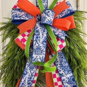 May include: A decorative bow featuring a mix of ribbons including a blue and white floral pattern, solid orange, bright lime green, and a pink and white checkered print. The bow is arranged with multiple loops and long trailing tails.