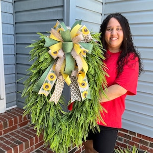 May include: A vibrant green wreath with a lush, leafy base. The wreath is adorned with a large bow featuring yellow sunflower ribbon, green and beige ribbons. The wreath is held by a person wearing a red shirt and black shorts.