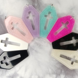 May include: Eight different colored coffin-shaped charms with a cross cutout. The charms are made of clear resin with glitter embedded in the resin. The charms are all different colors, including pink, purple, blue, green, black, and white.