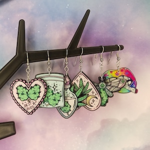 May include: Five pairs of dangle earrings with a cannabis leaf theme. The earrings are made of acrylic and feature various designs, including a heart with the text "Best Buds", a jar with a cannabis plant, a potion bottle with a cannabis plant, a crescent moon with a cannabis plant, and a butterfly with a cannabis leaf.