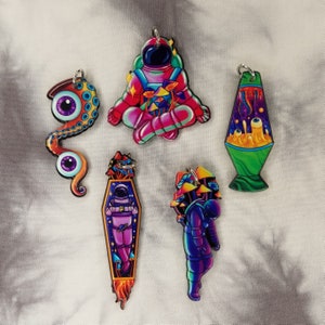 May include: Five colorful keychains featuring different designs. One keychain is a pink astronaut sitting cross-legged with mushrooms around them. Another keychain is a green lava lamp with a swirling orange and yellow design. A third keychain is a purple astronaut with mushrooms growing on their head. A fourth keychain is a purple and blue octopus with two large eyes. The final keychain is a purple astronaut in a rocket ship.