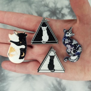 May include: Four black and white cat themed acrylic charms. One charm features two cats hugging, another features a cat sitting with a crescent moon, another features a cat sitting with a sun, and the last charm features a cat sitting with a starry sky.